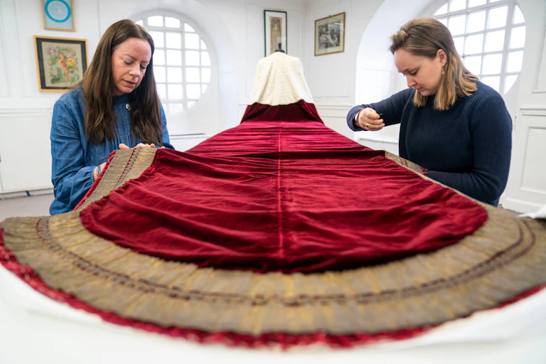 Two women work on a red velvet cape.