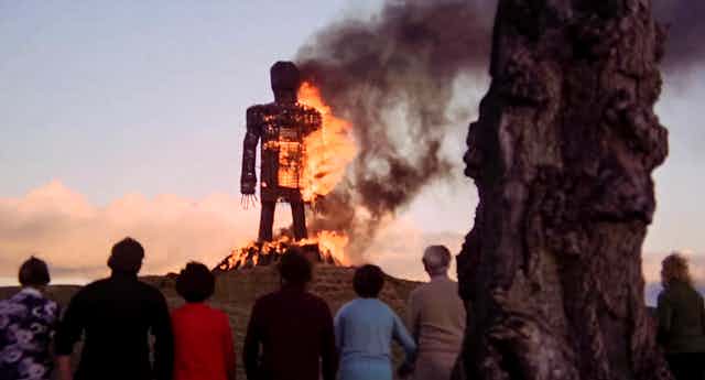 A giant straw effigy of a man on fire with some people looking on.