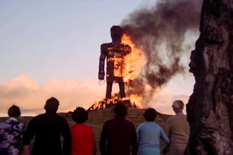 A giant straw effigy of a man on fire with some people looking on.