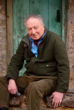 A middle aged man sitting on a doorstep wearing a shirt, cravat and cardigan.