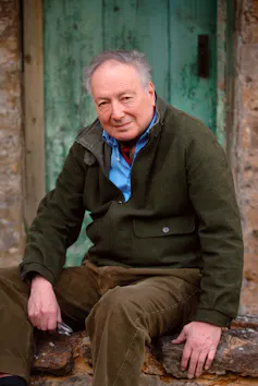 A middle aged man sitting on a doorstep wearing a shirt, cravat and cardigan.