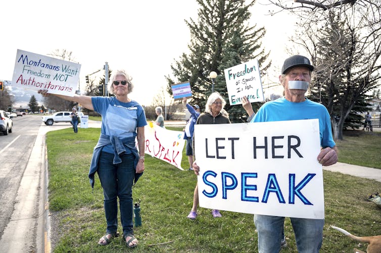 A few white people with grey hair wear blue tee shirts and hold up signs that say 'Montanans want freedom not authoritarians' and 'let her speak.' One man holding a sign has tape over his mouth.