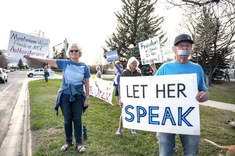 A few white people with grey hair wear blue tee shirts and hold up signs that say 'Montanans want freedom not authoritarians' and 'let her speak.' One man holding a sign has tape over his mouth.