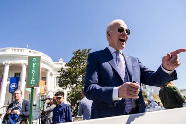 A white haired man in blue jacket and tie and aviator sunglasses pointing his finger and laughing.