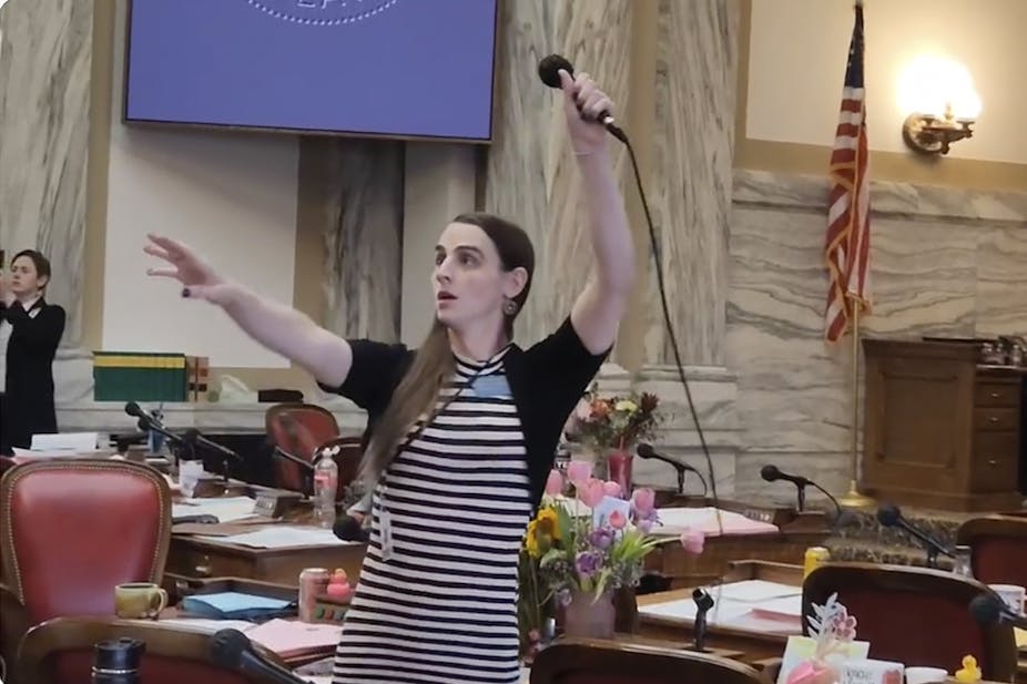 A woman wears a black and white striped dress and black cardigan, and raises a microphone in the air. She stands in a government looking room with an American flag and tables behind her. 