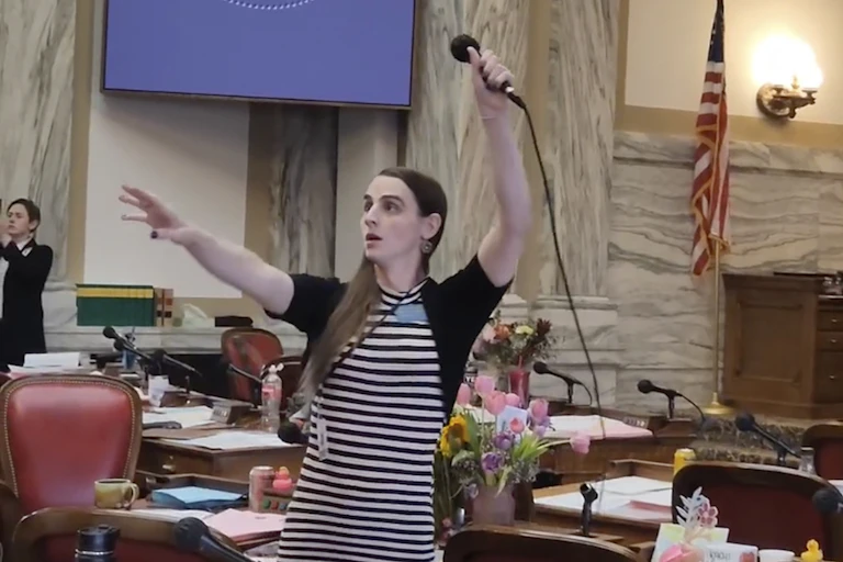 A woman wears a black and white striped dress and black cardigan, and raises a microphone in the air. She stands in a government looking room with an American flag and tables behind her.