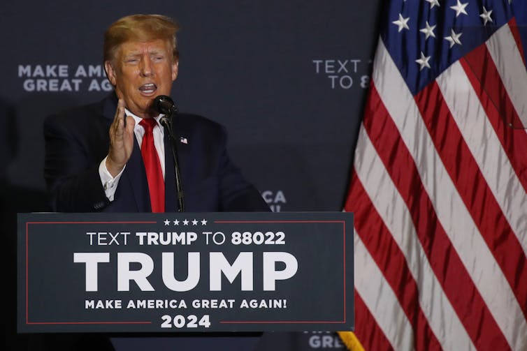 A man in a blue suit, red tie and white shirt, standing behind a Trump sign and next to a big American flag, gesturing with his right hand.