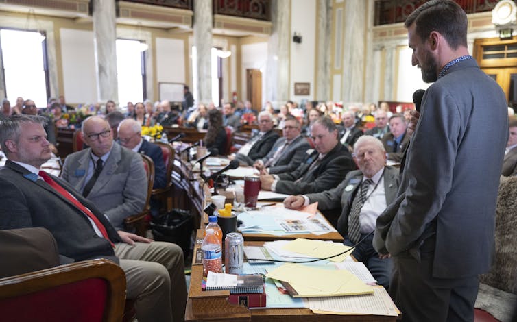 A large group of men in suits sit around tables with microphones.