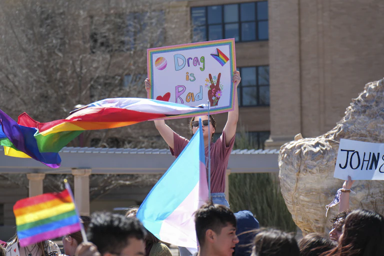 People at a protest waving rainbow flags, a woman carried a placard that reads: drag is rad.