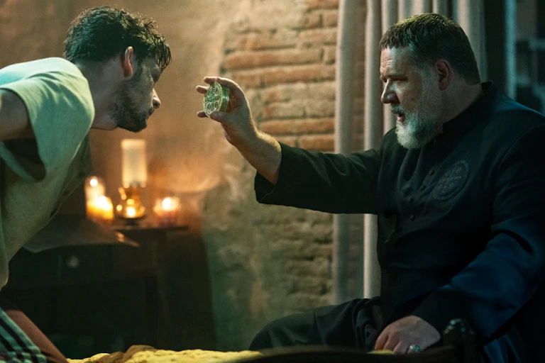 A man with a white beard, dressed in a priest robe, pointing a circular disc towards the forehead of another man.
