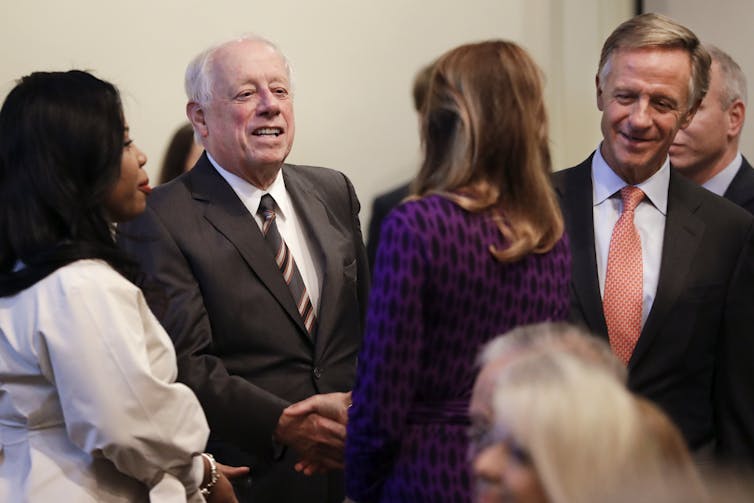 two white men are seen shaking hands and smiling with other people who's backs are turned