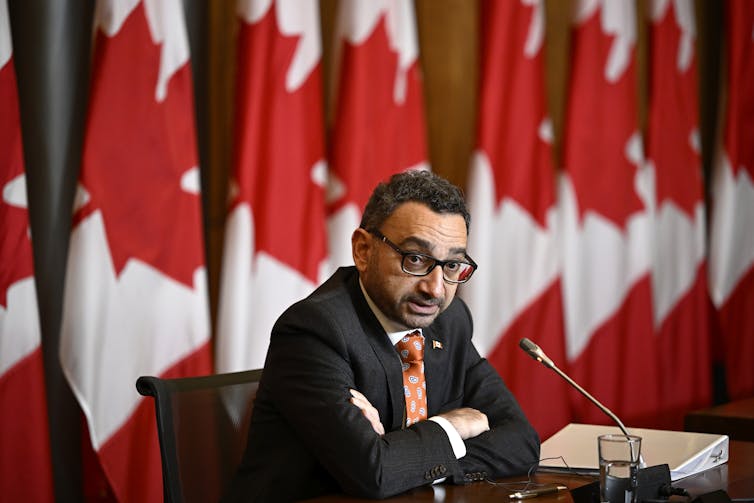 A man wearing a suit and glasses speaks from behind a microphone on a desk. A row of Canadian flags stand behind him.