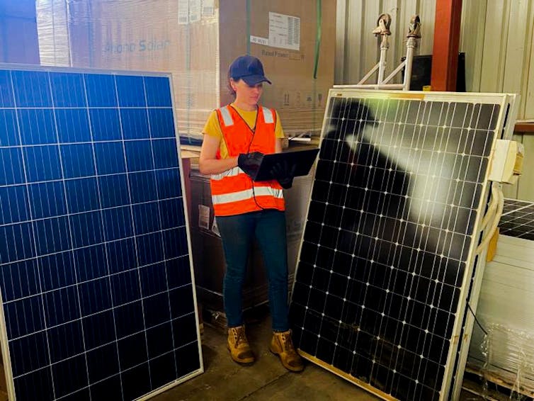 Young woman in hi-viz carries PV parts as she walks past two old solar panels