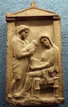 Un relief sculpté montre un homme debout tenant un enfant emmailloté, avec une femme assise à côté d’eux