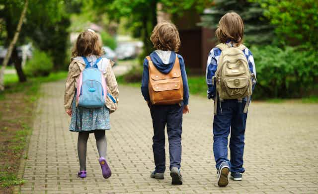 Three children in backpacks walk across the pavement