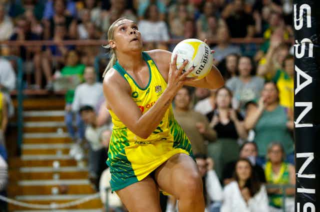 Indigenous woman in green and gold uniform taking a shot with a netball in a crowded stadium