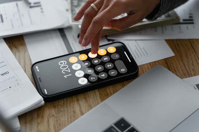 A hand uses the calculator on a smart phone, surrounded by papers.
