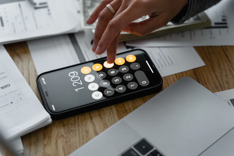 A hand uses the calculator on a smart phone, surrounded by papers.