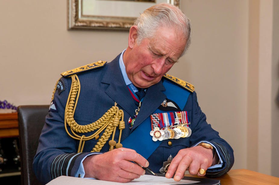 King Charles III in military dress, sits at a desk and signs a document