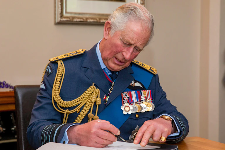 King Charles III in military dress, sits at a desk and signs a document