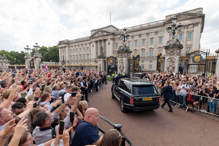 A black car arrives at Buckingham Palace while hundreds of onlookers wave