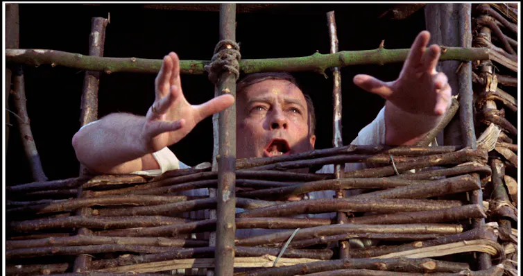 A man imprisoned in a makeshift prison made of wood, his hands outstretched in plea.