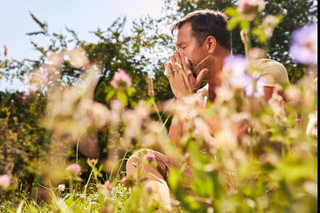 A man sitting in a flowering garden blows his nose.