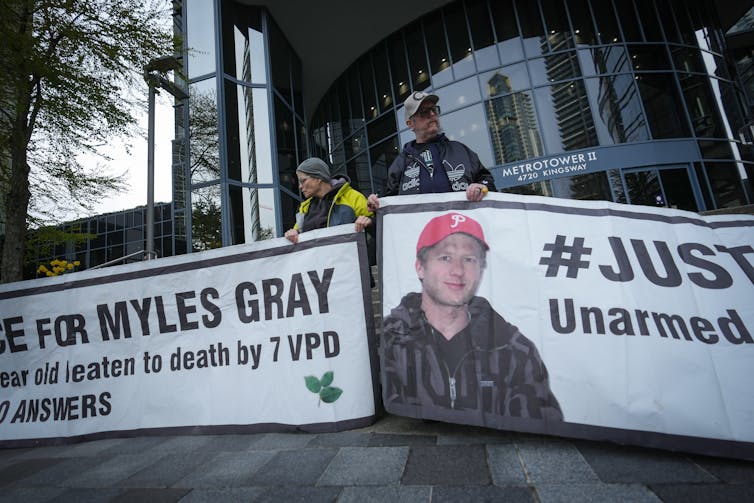 Protesters hold banners with a photograph of a man in a ski jacket and red ball cap that says Justice for Myles Gray.