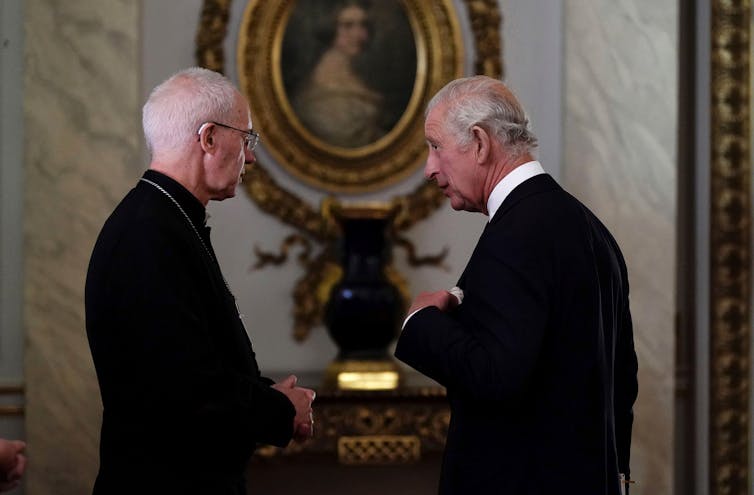 The Archbishop of Canterbury and King Charles, both wearing black, speak to each other in front of an ornate vase on a side table in Buckingham Palace