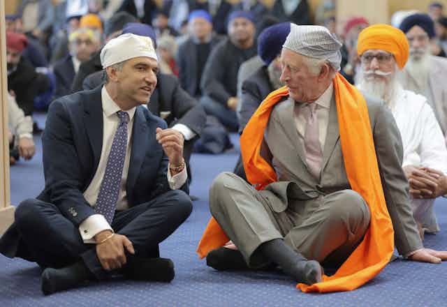 King Charles, wearing a Sikh head covering and scarf, sits on the floor surrounded by other Sikh men.