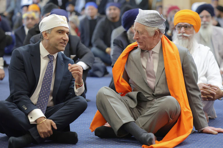 King Charles, wearing a Sikh head covering and scarf, sits on the floor surrounded by other Sikh men.