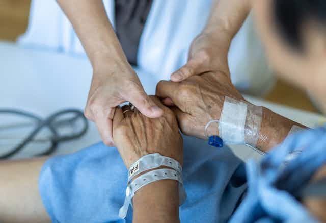 An older person's hand with a hospital bracelet on the wrist and the hands of a younger person out of frame wearing a white coat