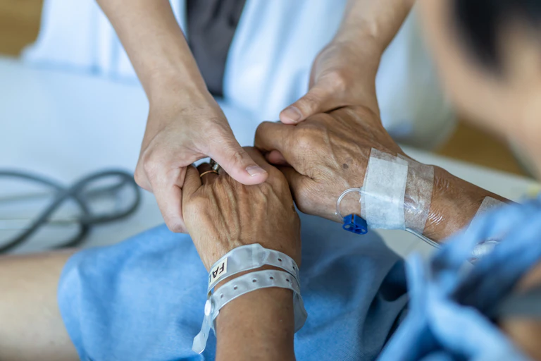 An older person's hand with a hospital bracelet on the wrist and the hands of a younger person out of frame wearing a white coat