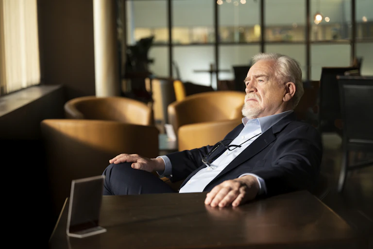 Brian Cox reclining in a chair, wearing a navy suit.