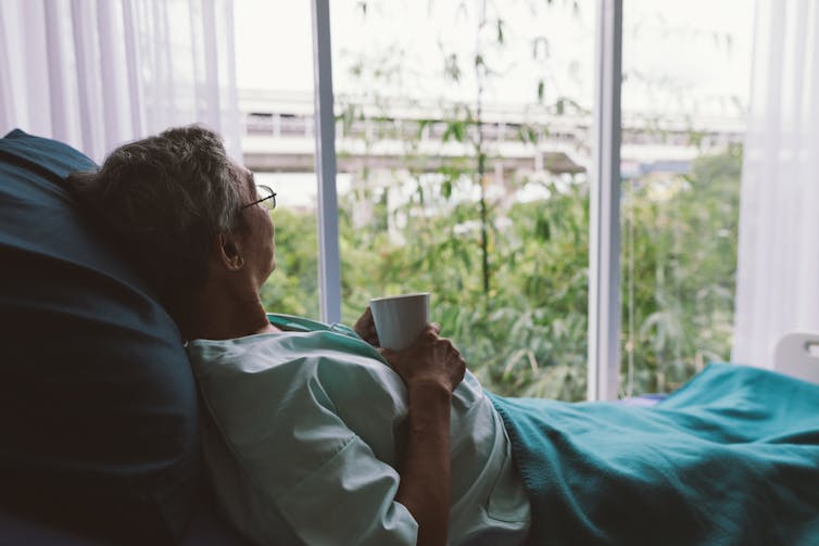 A person in a hospital bed looking out a large window with a mug in their hands.