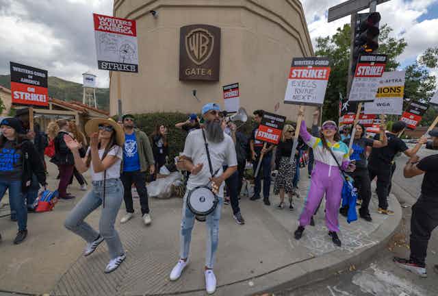Striking workers holding signs, chanting and dancing in front of a movie studio.