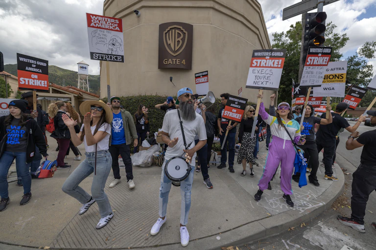 Striking workers holding signs, chanting and dancing in front of a movie studio.