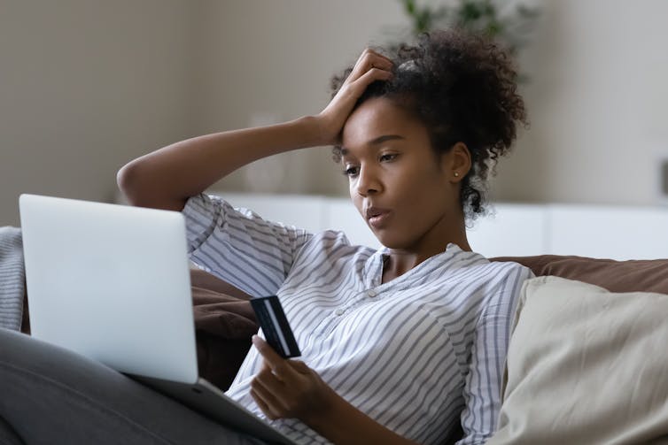 A worried-looking young woman sitting on the couch with a credit card in one hand and her head resting on her other hand. she is looking at a laptop computer.