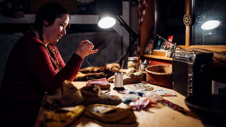 A woman sitting at a work desk doing beadwork.