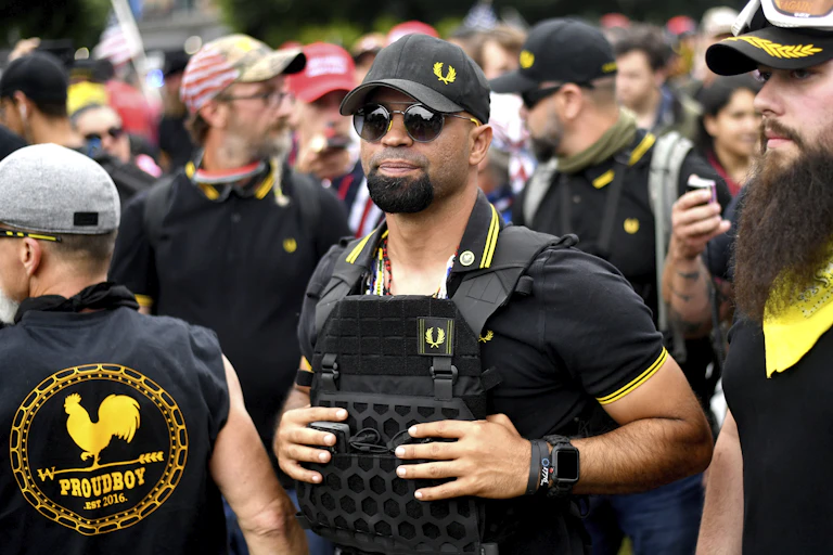 A person wearing a tactical vest stands in a crowd.