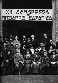 A black and white formal portrait of three rows of men and women in coats and suits posing in front of a banner.