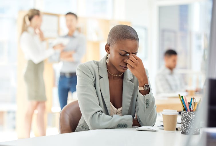 Woman at desk with her hand on her face/headache, people in suits talking in the background.