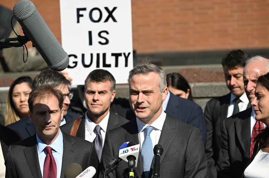 Un homme parle devant des micros, derrière lui un panneau sur lequel il est écrit « Fox is guilty »