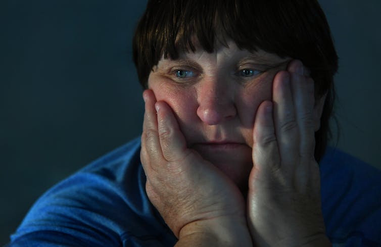 A white woman with short brown hair with her hands holding her face looks sad.