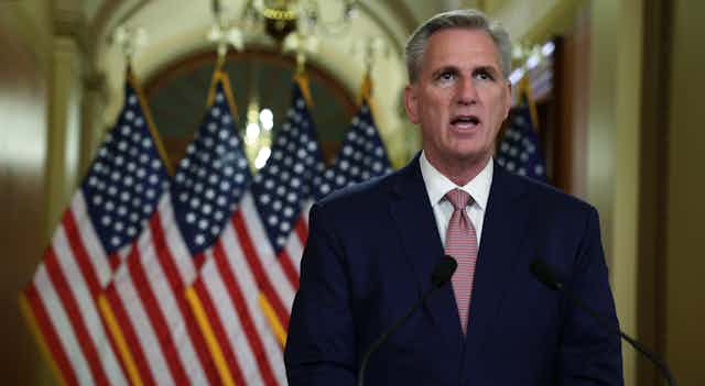A white man with gray hair wearing a mauve tie and blue blazer speaks in front of four American flags.