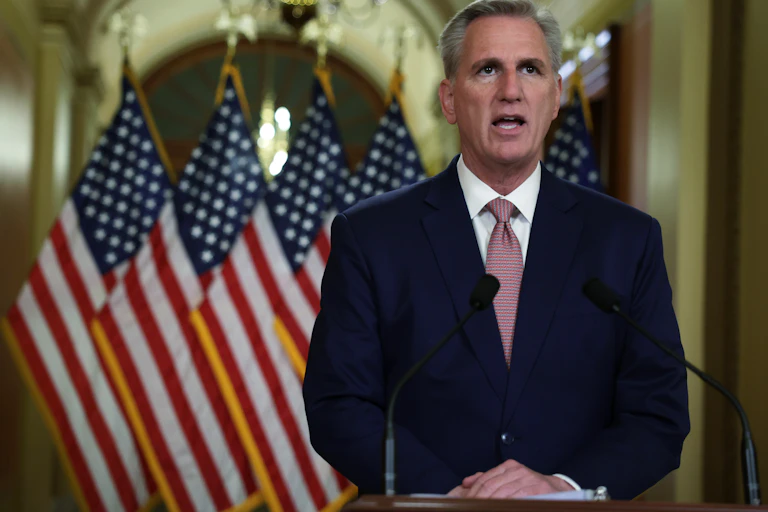 A white man with gray hair wearing a mauve tie and blue blazer speaks in front of four American flags.
