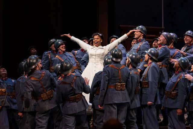A woman sings on a stage in a white dress, beaming with her arms in the air. All around her are soldiers in uniform, singing too.