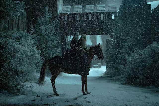 Un hombre y una mujer montan en caballo en un pueblo de noche mientras nieva.