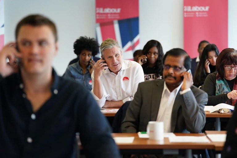 Keir Starmer on the phone in the middle of rows of tables at which other people are sitting, also speaking on phones.