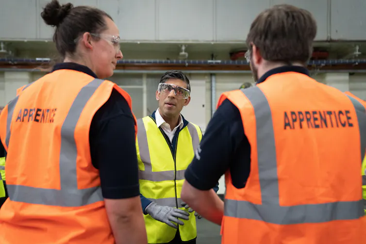 Rishi Sunak wearing a high viz jacket talking to two people wearing orange jackets with the word 'apprentice' on the back.